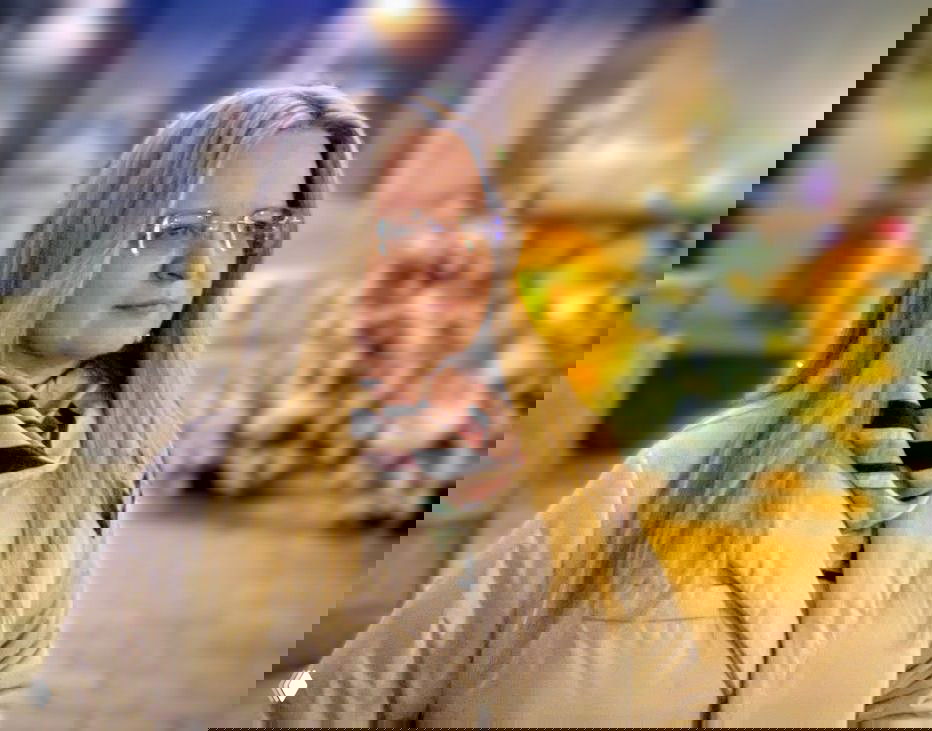 Портрет женщины в светлой зимней куртке и шарфе, в очках; на размытом фоне видно уличную подсветку и декоративные ели.