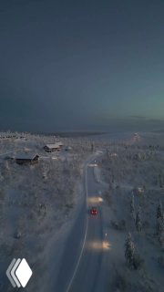 Заснеженный вечерний пейзаж Лапландии: домики, покрытые снегом, хвойный лес и заснеженная дорога с проезжающими автомобилями.