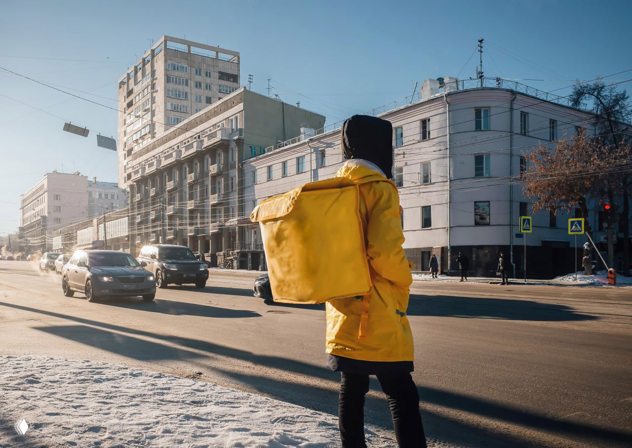Курьер в жёлтой куртке и с большой доставочной сумкой стоит у перекрёстка на фоне машин и зданий в солнечный зимний день.