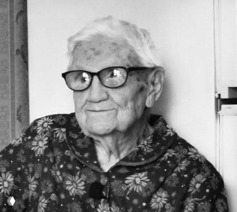 Чёрно‑белая фотография пожилой женщины в очках и цветочном халате — портрет Клавдии Михайловны Гадючкиной в домашнем интерьере, видна лёгкая улыбка и возрастные черты.