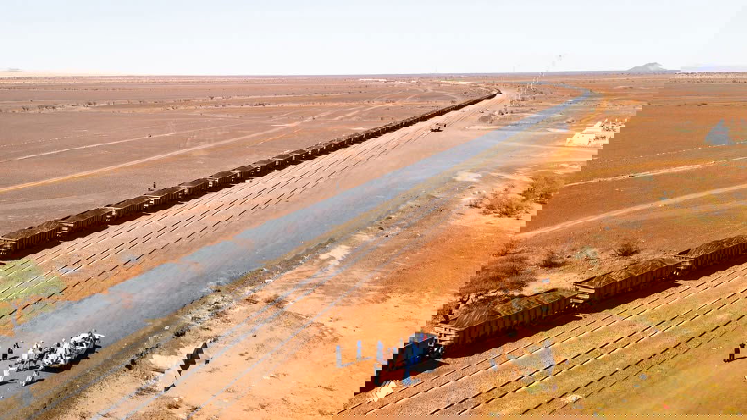 Вид сверху на длинную цепь вагонов Iron Ore Train в пустынном ландшафте Мавритании, горы на горизонте и отдельные деревья вдоль пути