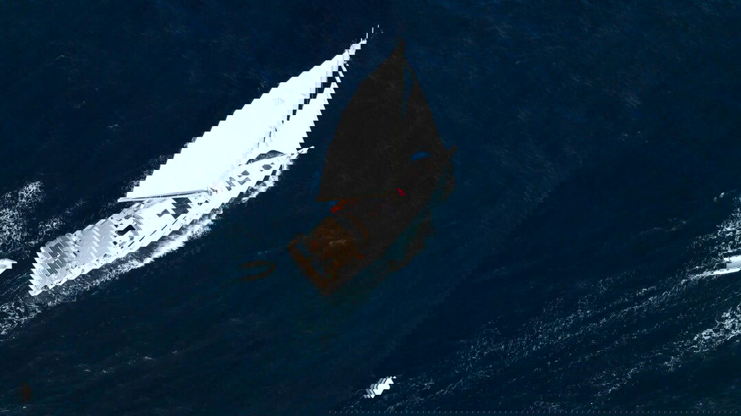 Вид сверху на парусную яхту и буксируемую надувную лодку; кадр с высоты показывает конфигурацию палубы и волны вокруг судна.