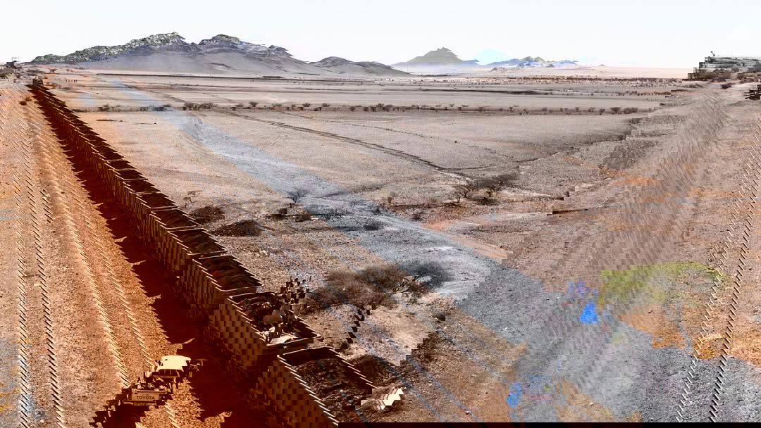 Аэрофотоснимок поезда с открытыми вагонами, заполненными чёрной железной рудой, на фоне сухой пустынной ровнины и гор