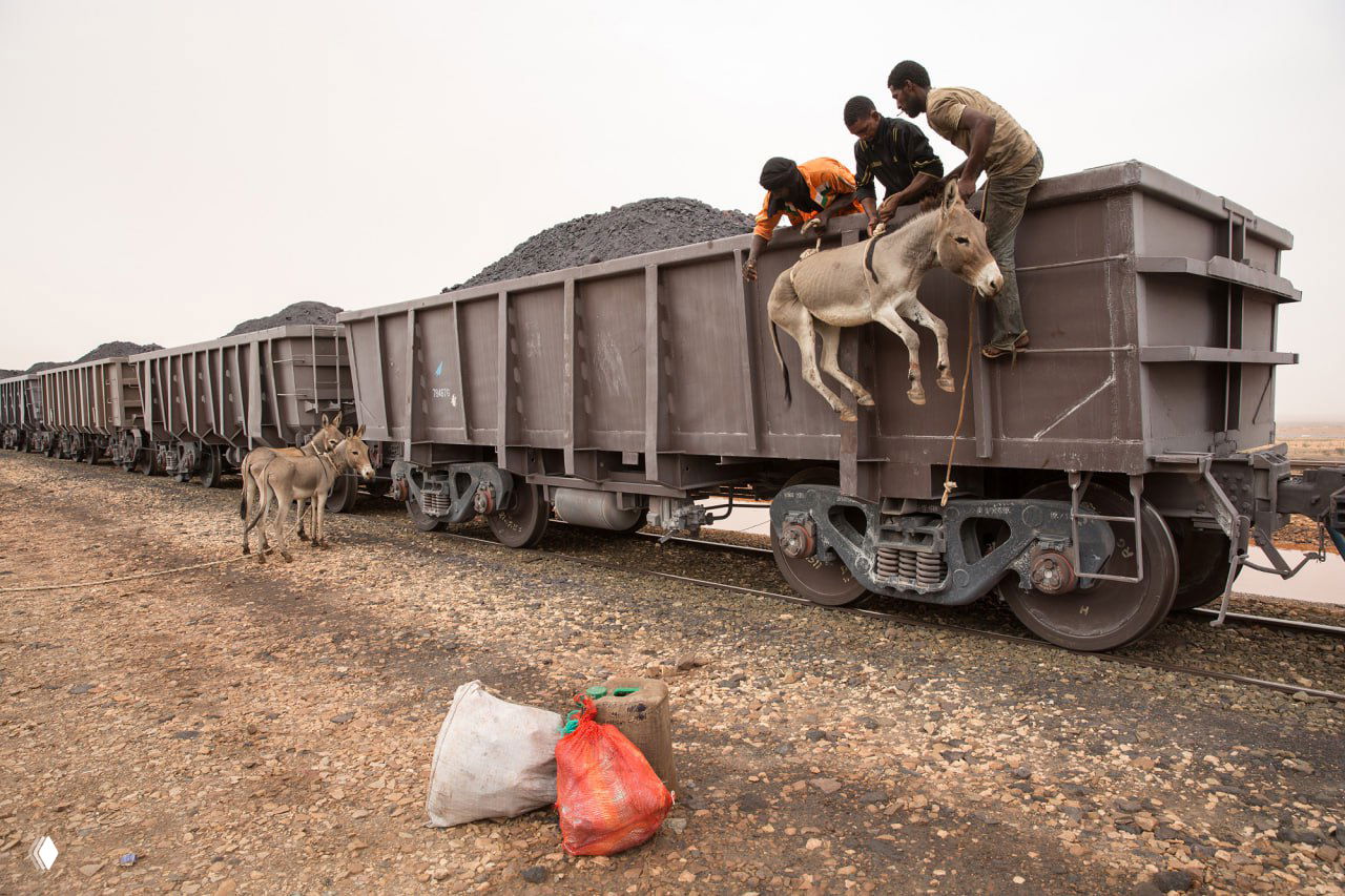 Местные жители и ослы поднимаются на борт рудного вагона: люди часто путешествуют, сидя прямо на руде, в суровых температурных условиях пустыни