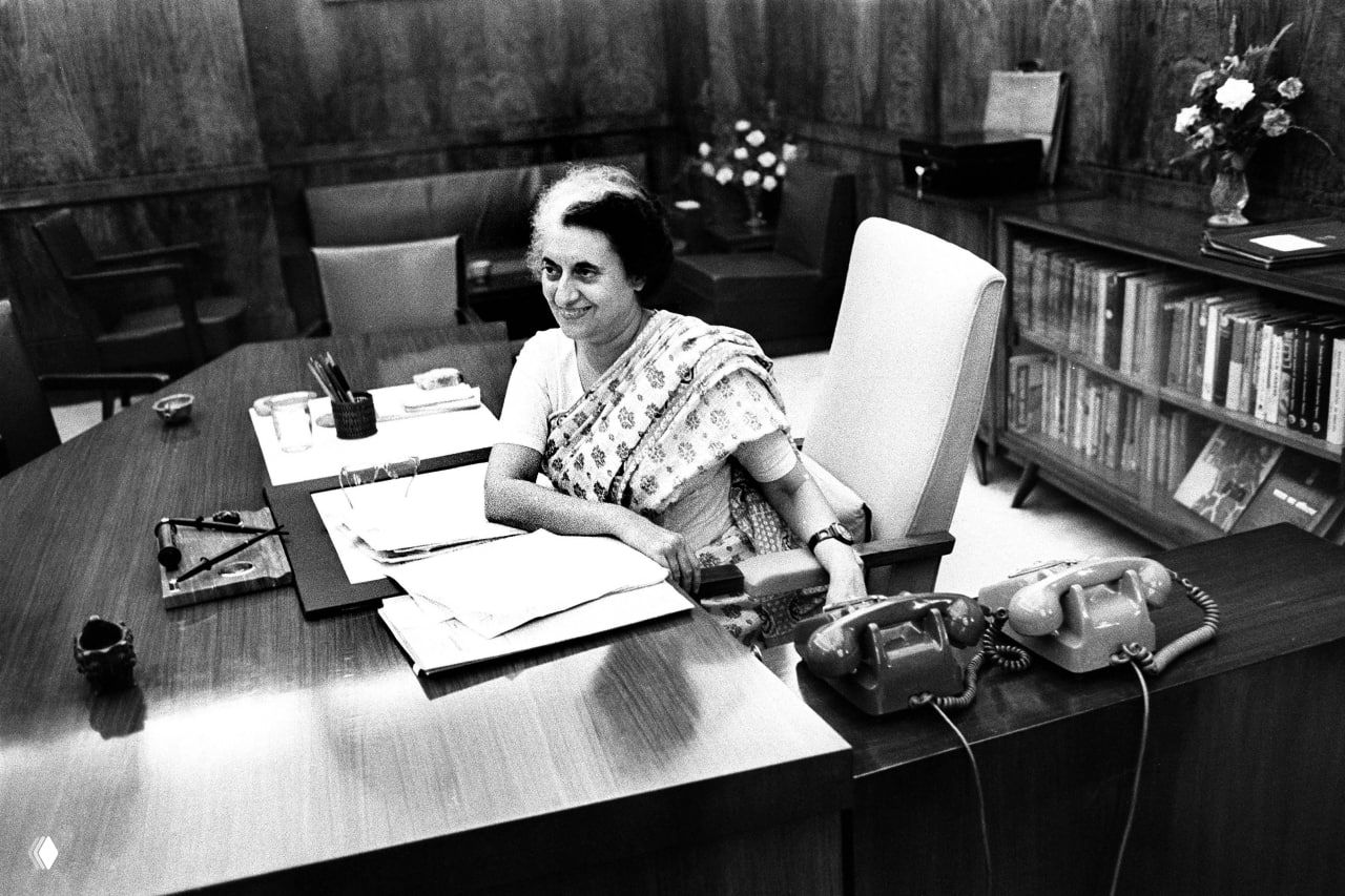Чёрно-белая архивная фотография женщины-политика за рабочим столом с документами и двумя телефонами, официальный кабинет.