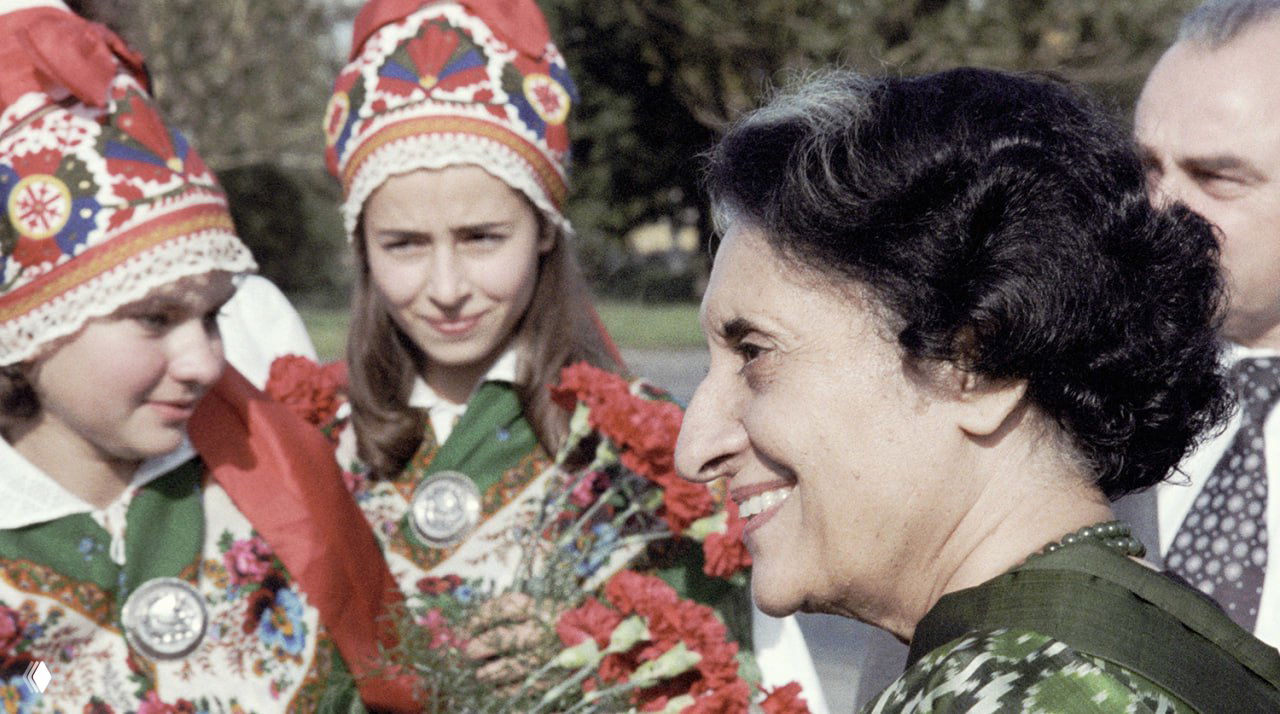 Цветная фотография женщины в профиль на встрече с молодыми людьми в национальных костюмах, вручение цветов и приветствие.