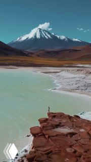 Панорамный вид ландшафта пустыни Атакама: солёное озеро на переднем плане и заснеженный вулкан на далёком горизонте, пустынная равнина.