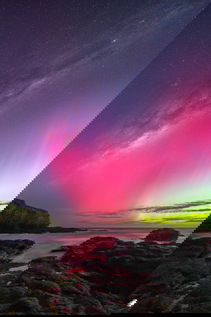 Ночное прибрежное фото: скалистый берег и океан на переднем плане, над ними Млечный Путь и яркие розово‑зеленые полосы сияния в небе.
