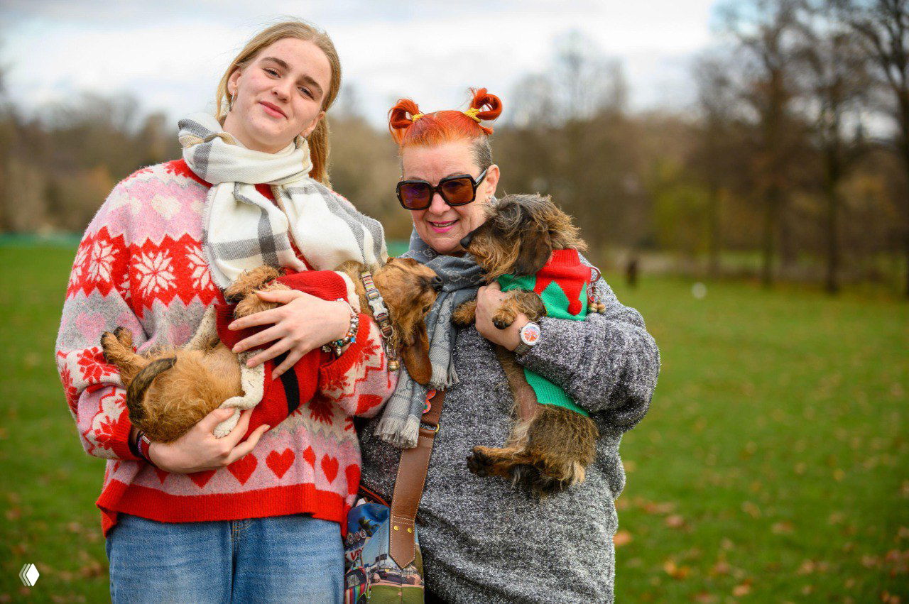 Две женщины держат на руках нескольких такс в праздничных одеждах на зелёной поляне Гайд‑парка — участники и гости фестиваля.