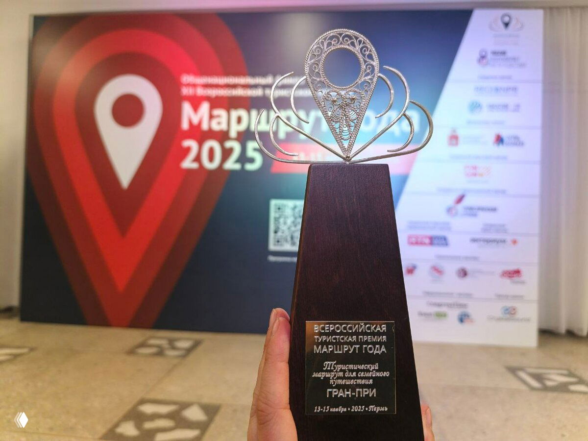 Крупный план статуэтки и кубка премии «Маршрут года» на фоне баннера мероприятия с логотипом и датами.