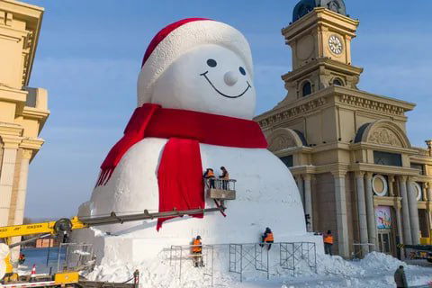 Рабочие на подъемнике отделывают лицо и шарф большого снеговика в Харбине: вид крупным планом, красный шарф и улыбка на снежной фигуре.