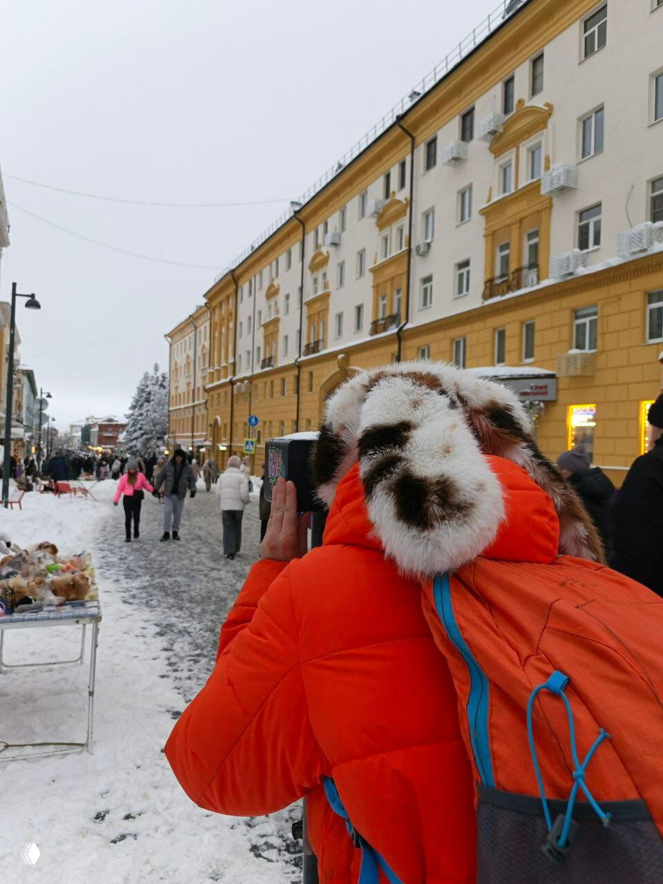 Сзади снятая путешественница в оранжевой куртке и меховой шапке фотографирует уличную ярмарку на снежной городской улице с исторической застройкой.