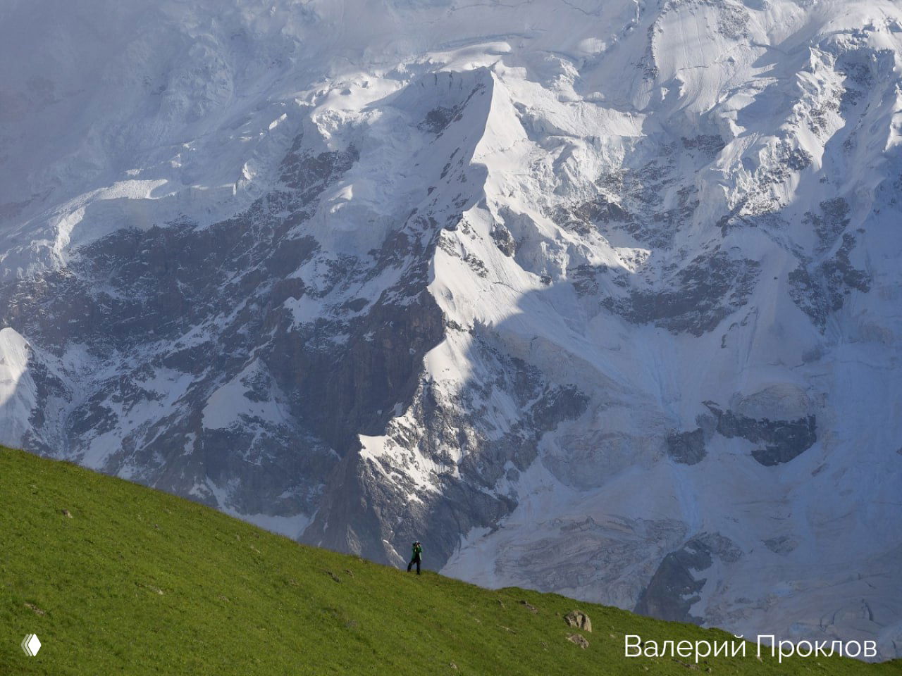 Альпинист на зелёном склоне на фоне огромной заснеженной горной стены — контраст масштаба человека и кавказских пяти‑тысячников.