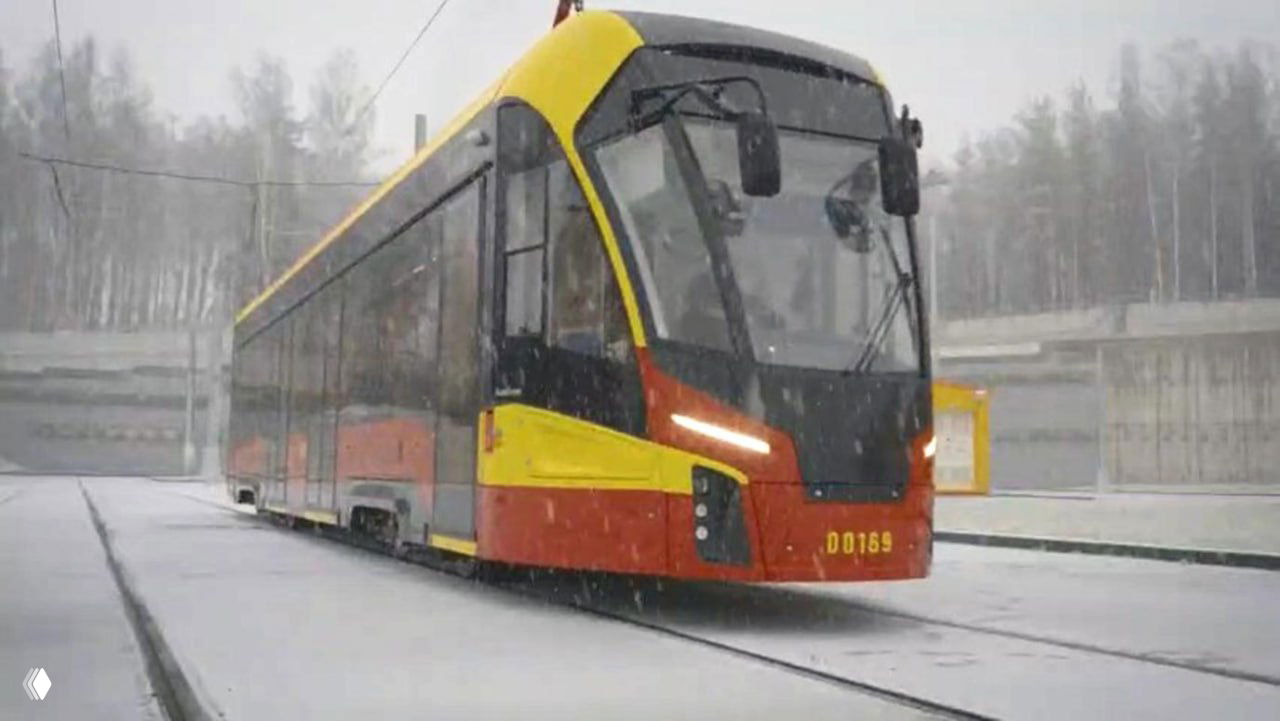 Беспилотный трамвай «Львенок» красно‑жёлтой окраски движется по линии в снежную погоду, видна платформа и деревья на заднем плане.