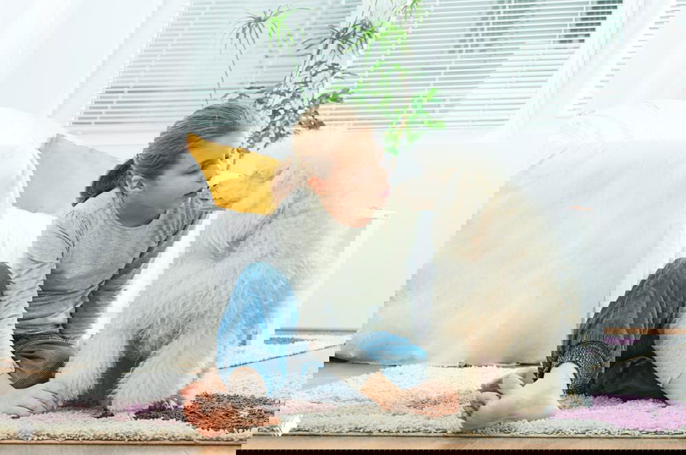 Девочка сидит на ковре в светлой квартире и нежно общается с большим белым пуделем — спокойная домашняя сцена контакта ребёнка и собаки.