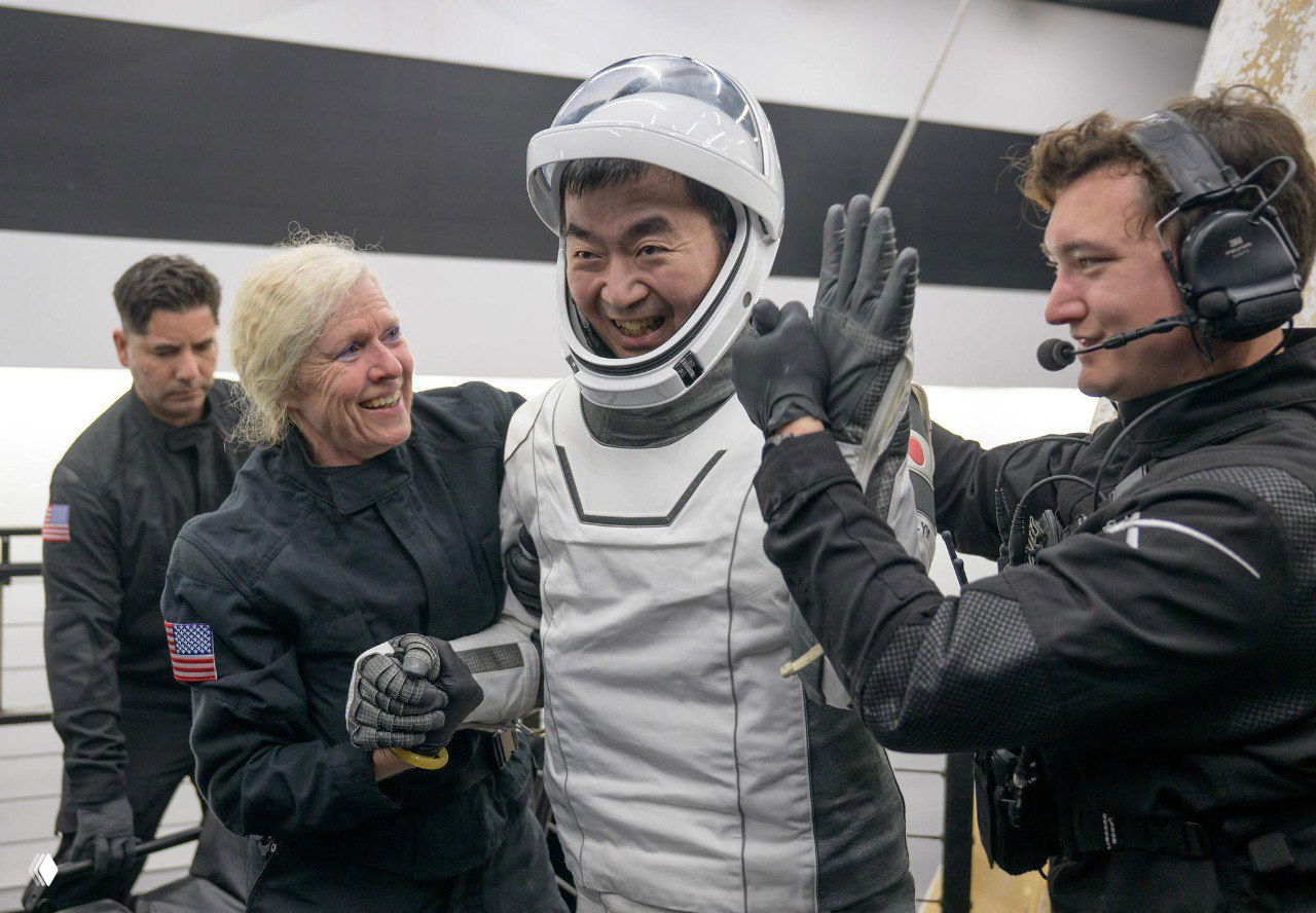 Астронавт азиатского происхождения в скафандре улыбается и получает помощь от персонала рядом с обгоревшей капсулой Crew Dragon внутри ангара.