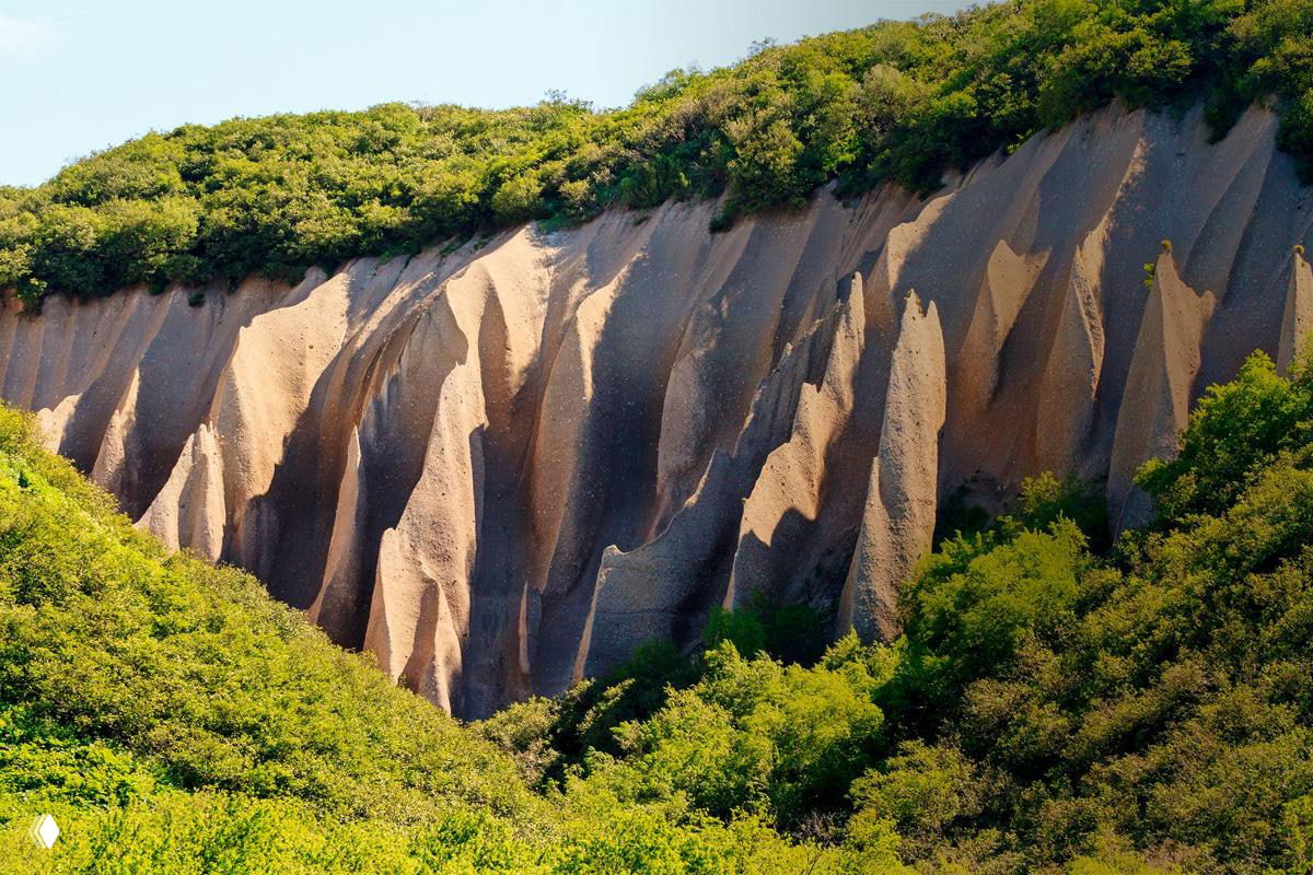 Панорамный вид на извилистые пемзовые стенки Кутхиных Бат, окаймлённые густой зеленью, солнечный свет подчёркивает фактуру и рельеф.