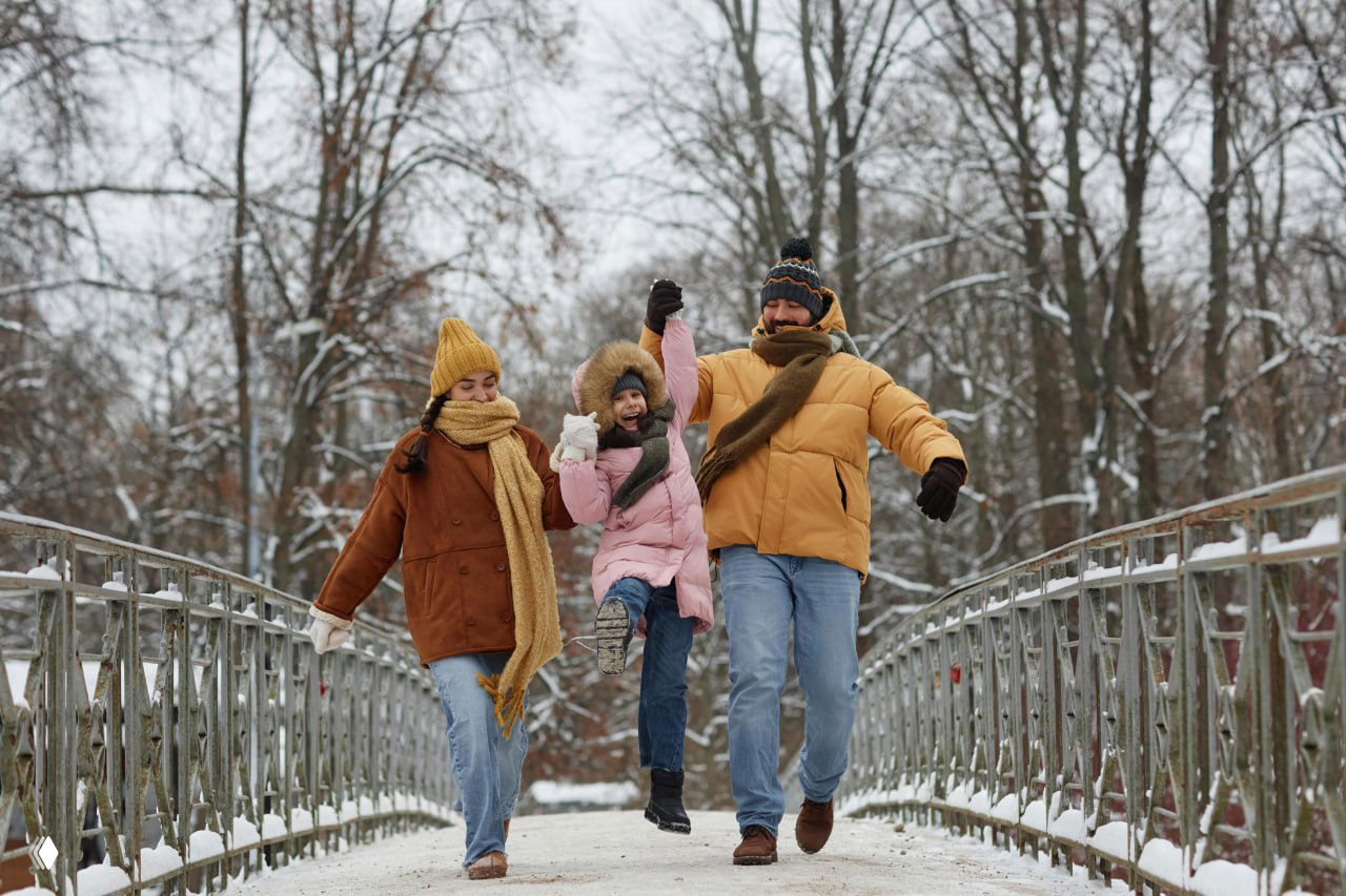 Семья в зимней одежде (мама, папа и ребенок) идет по заснеженному мосту в городском парке, праздничная и дружная зимняя сцена.