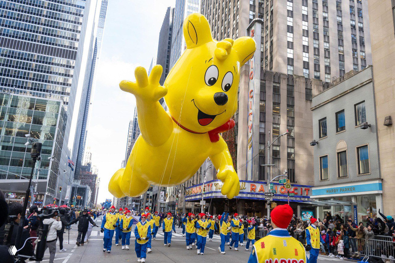 Огромный жёлтый надувной шар в виде мультгероя над Манхэттеном на параде Macy’s; видны зрители, помощники и городские фасады вдоль маршрута.