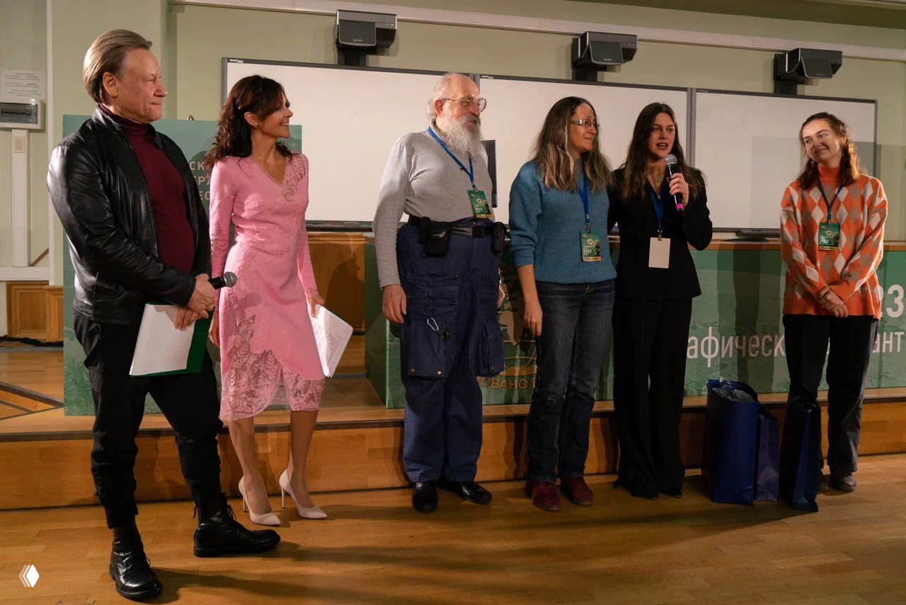 Несколько организаторов и участников стоят на сцене перед залом; видна эмблема мероприятия и оформление сцены.