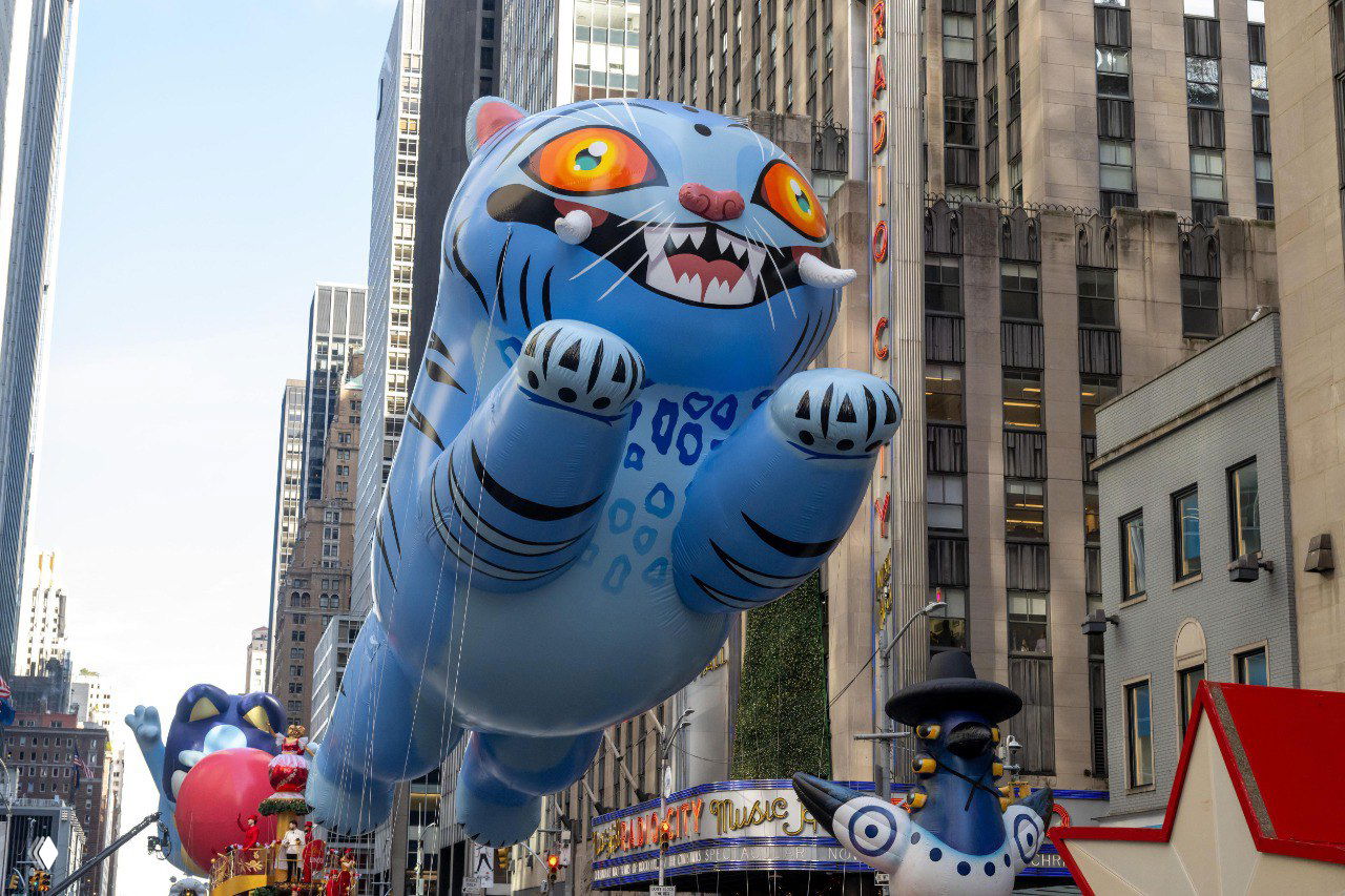 Голубой надувной кот с большими глазами летит над Пятой авеню в Нью-Йорке; на заднем плане видны другие шаровые фигуры и высотки.