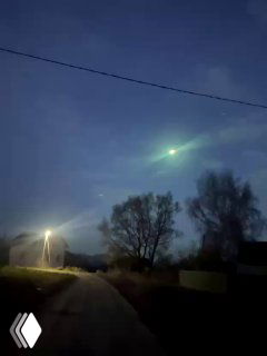 Ночное фото болида над деревней: зелёное светящееся тело в небе над дорогой, видны деревья и уличный фонарь, кадр очевидца.