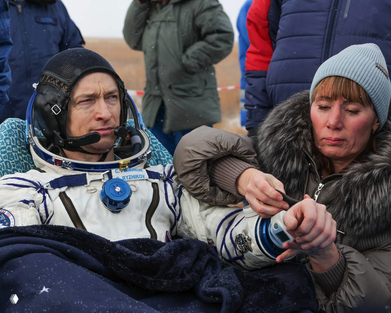 Космонавт Сергей Рыжиков после посадки на степи: в скафандре, рядом помогают с оборудованием и медицинскими проверками, заметна холодная погода.