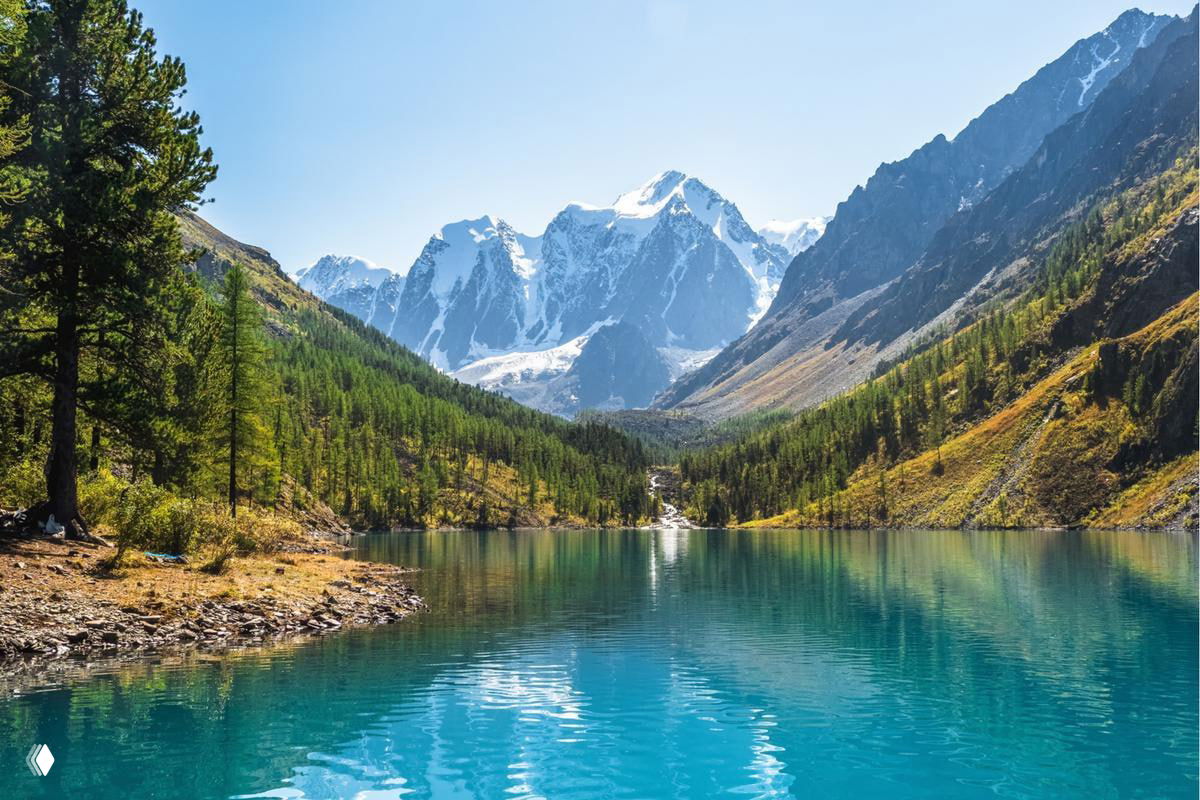 Шавлинское озеро с зеркальной, прозрачной поверхностью на фоне заснеженных вершин и хвойных лесов в высокогорной долине Алтая на высоте около 2000 м.