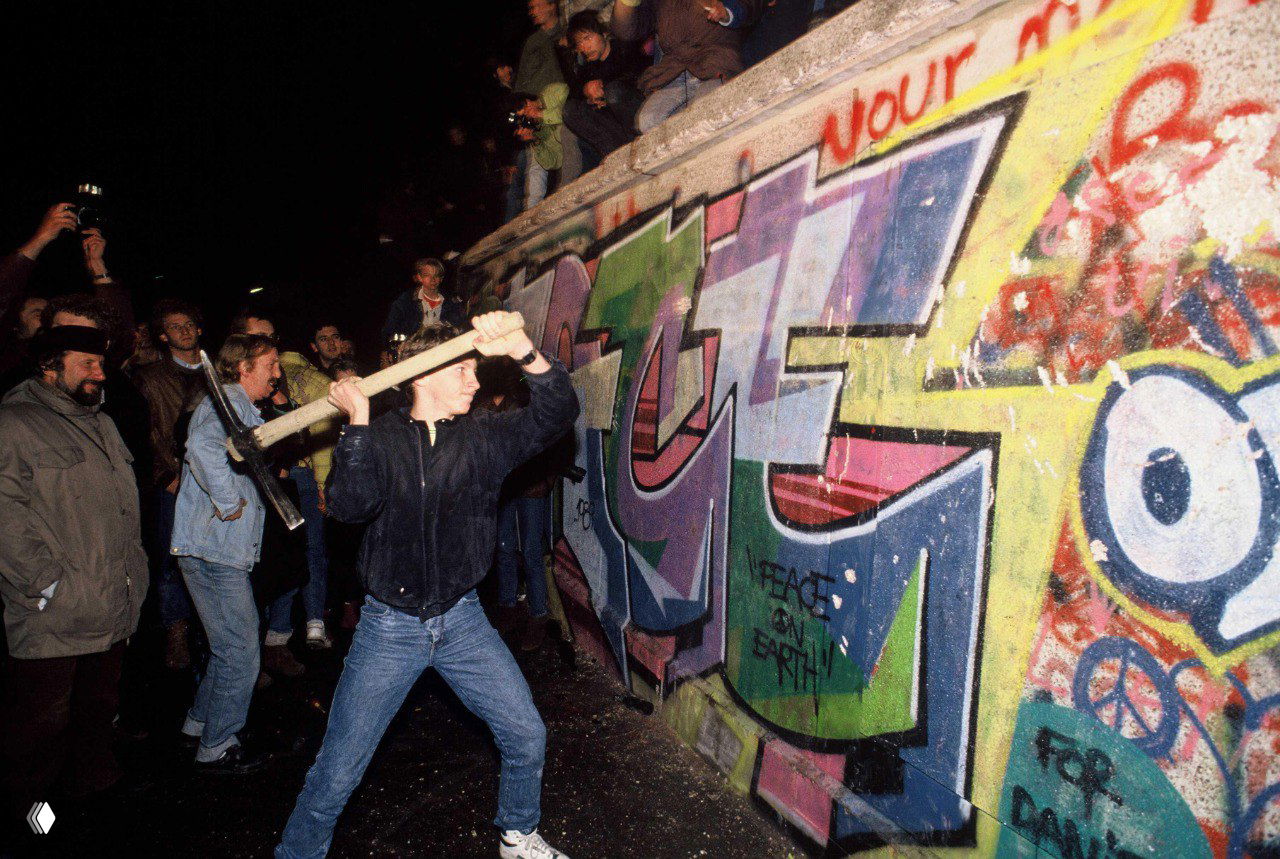 Ночное изображение: мужчина с кувалдой и ломом бьёт часть Берлинской стены, вокруг толпа, видны цветные граффити и бурная атмосферa протеста и радости.