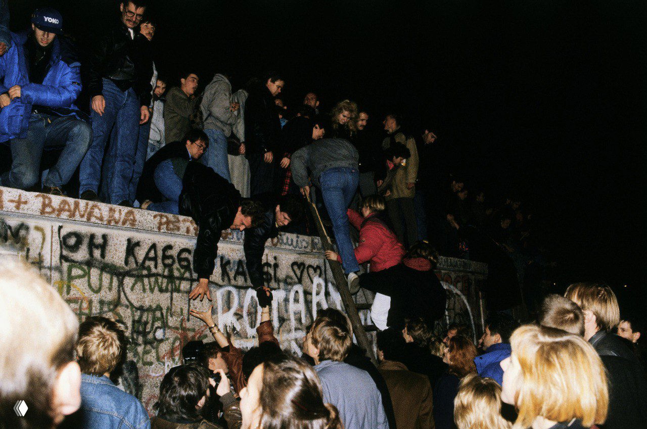 Люди карабкаются на Берлинскую стену и помогают друг другу перелезть, кадр ночного массового участия горожан в демонтаже барьера и объединении городского пространства.