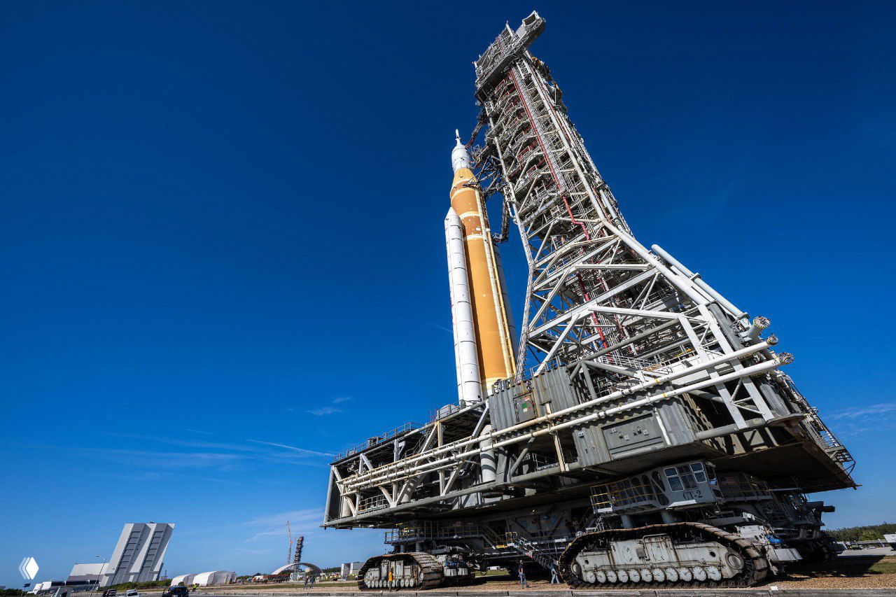 Крупный план нижней части ракеты SLS и массивной опорной тележки перед стартовой башней, металлическая конструкция на фоне синего неба.