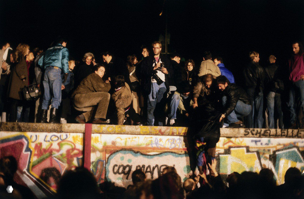 Камера фиксирует людей на стене и у её подножия ночью; на переднем плане — группа с фотоаппаратами, в центре — оживлённые действия активных участников событий 9 ноября 1989 года.