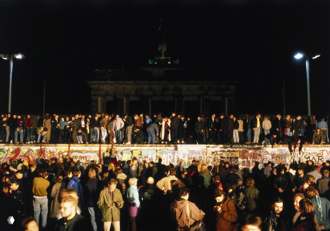 Широкая панорама: множество людей вокруг и на верхней части Берлинской стены ночью, вдалеке видны контуры Бранденбургских ворот и ночное освещение исторического места.