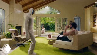 NEO в гостиной рядом с креслом и человеком: гуманоидный домашний робот в мягком вязаном корпусе, демонстрация взаимодействия в интерьере.