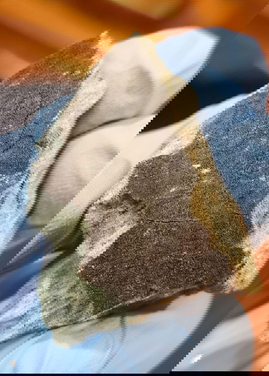 Крупный план фрагмента каменного метеорита в синих перчатках; видна тёмная поверхность с трещинами и светлая матрица, лабораторный фон.