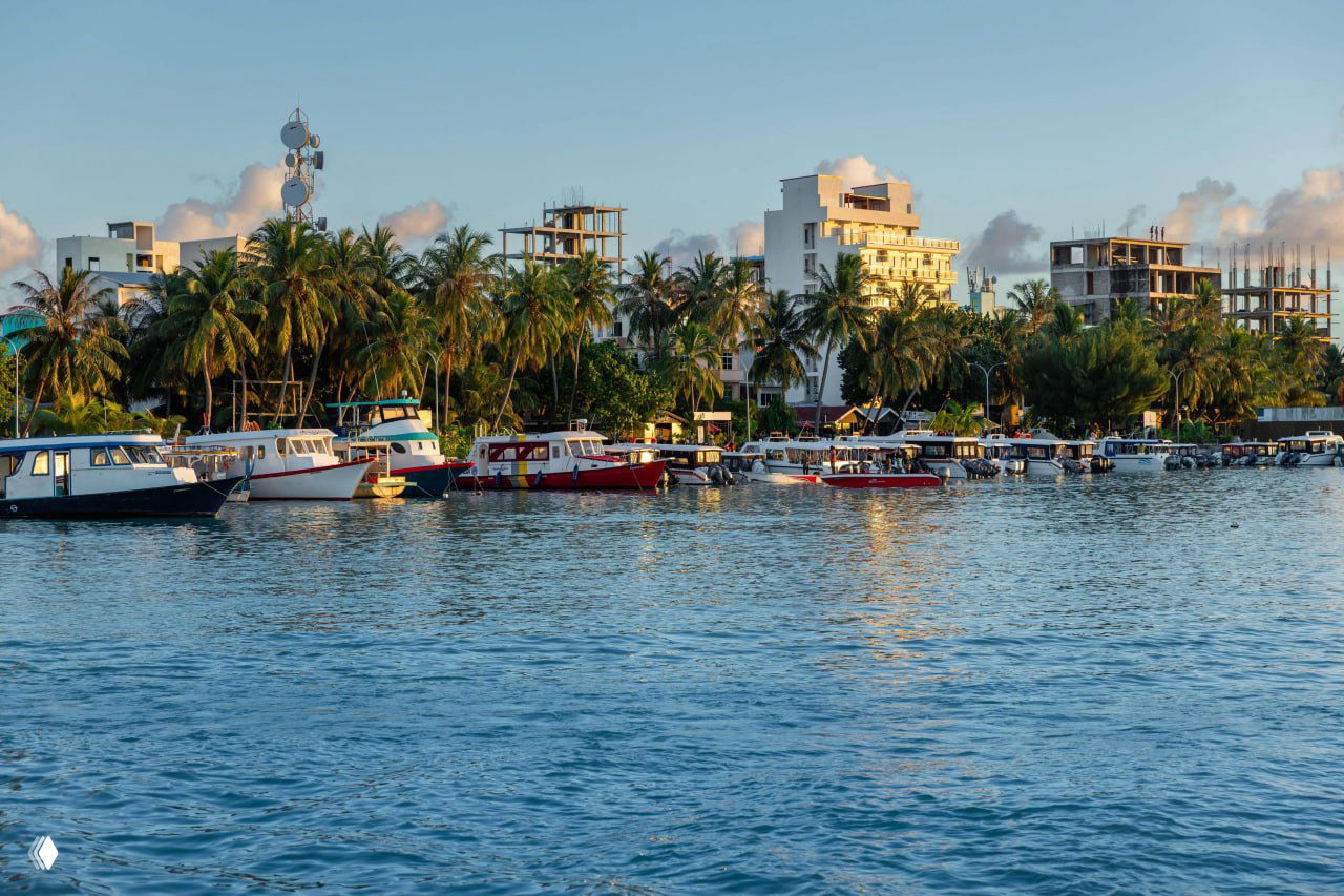 Лодки и катера у причала на Мальдивах на фоне пальм и прибрежных зданий; тёплый свет заката отражается в спокойной воде.