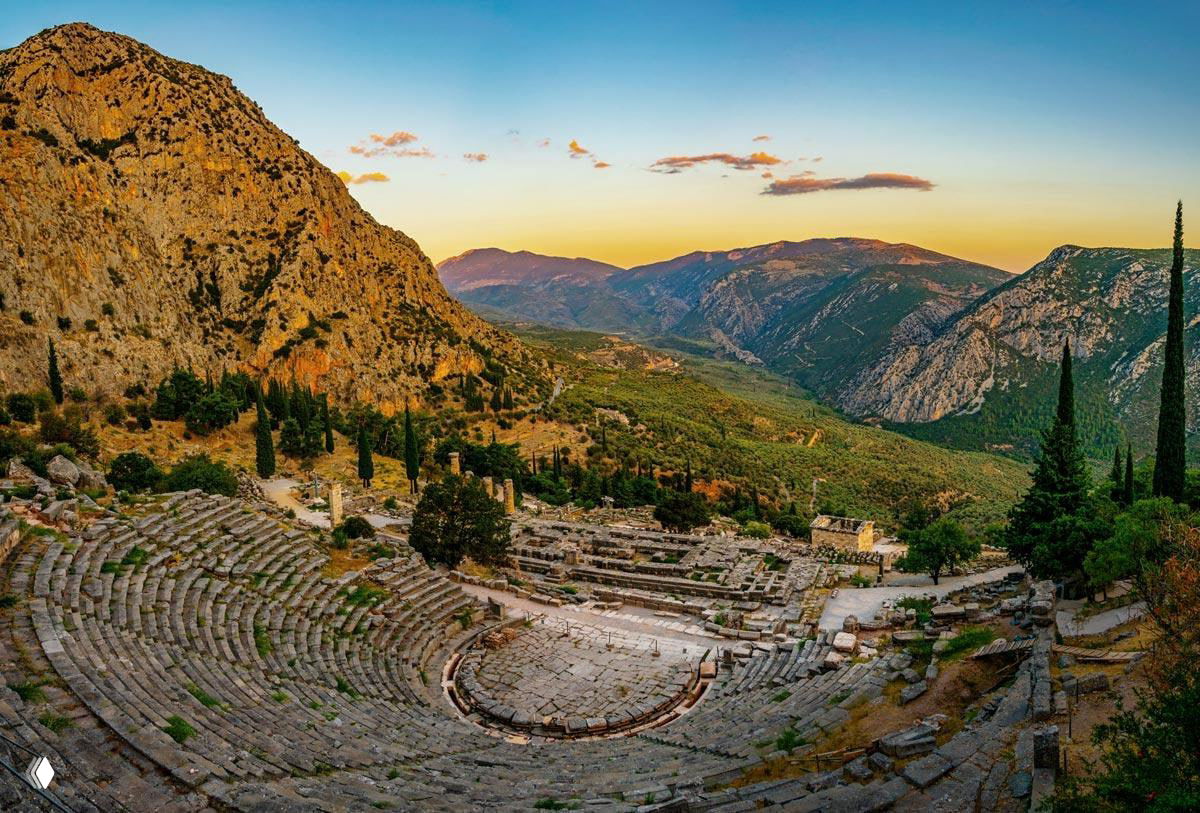 Древний театр на склоне горы с каменными трибунами и круглой оркестровой зоной, открывающий вид на долину и закатное небо.