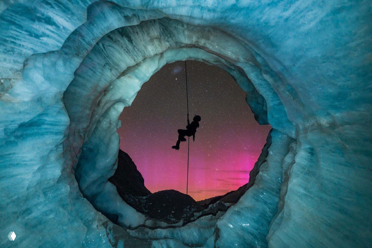 Силуэт альпиниста на спуске в ледяной пещере, вид на розово‑зеленые полярные сияния и звёздное небо через круглой формы отверстие в льду.