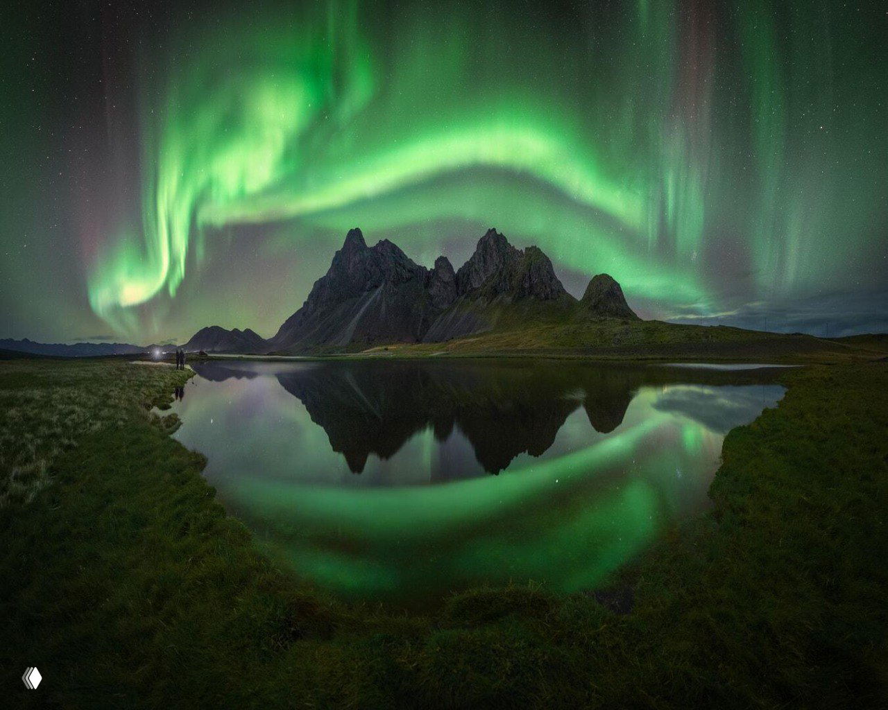 Горный пейзаж с зеркальным озером, над которым разворачивается широкая изумрудная лента полярного сияния, отражающаяся в воде.