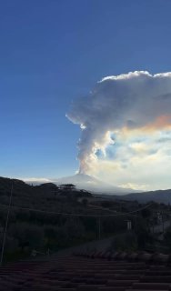 Вертикальное фото/видео: высокий столб пепла и газа от Этны, поднимающийся в небо над холмами и крышами домов на переднем плане.