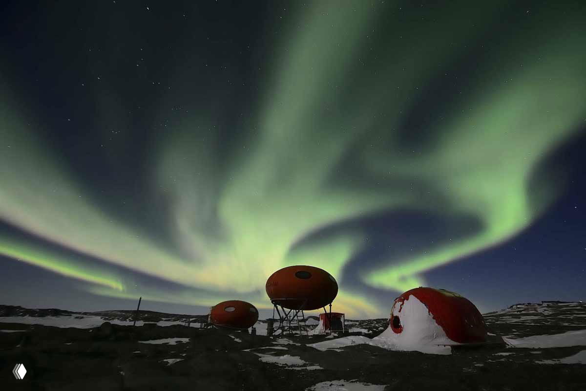 Яркое полярное сияние над антарктической базой с оранжевыми купольными модулями на снегу, зелёные ленты в небе.