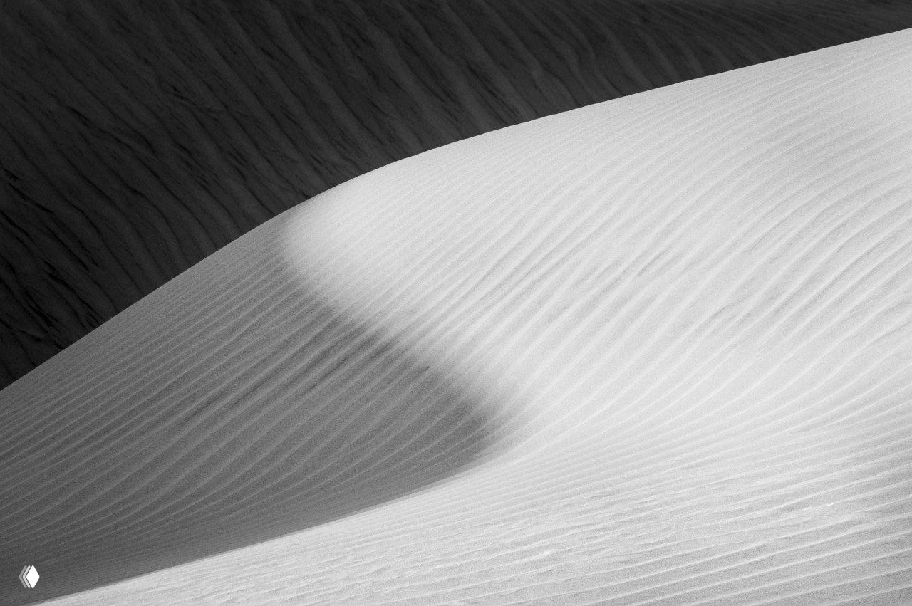 Черно‑белая фотография песчаной дюны с ритмическими текстурами и контрастом света и тени, графичный минимализм в природе.