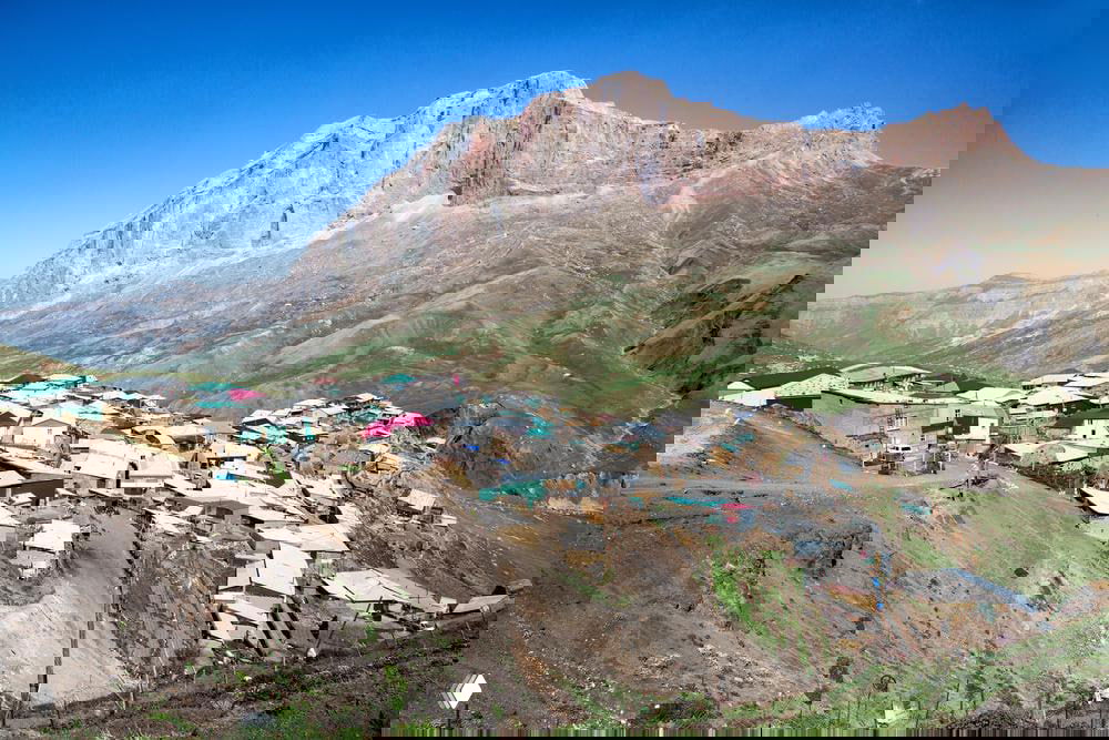 Панорамный вид на высокогорное село Куруш в Дагестане: дома на крутом склоне у подножия массивной скалы Базардюзю под ясным небом.