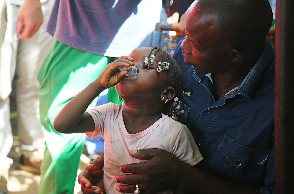 Маленькая девочка в африканской деревне принимает лекарство из стаканчика под присмотром взрослого — раздача антибиотика.