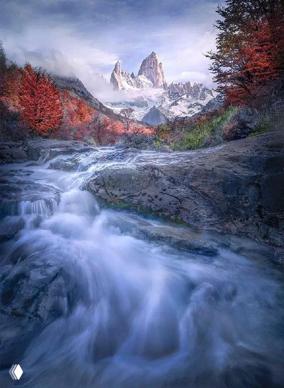 Ручей в горах на переднем плане и острые скалы с лиственным лесом в осенних тонах; длинная выдержка создаёт эффект шёлковой воды в кадре.