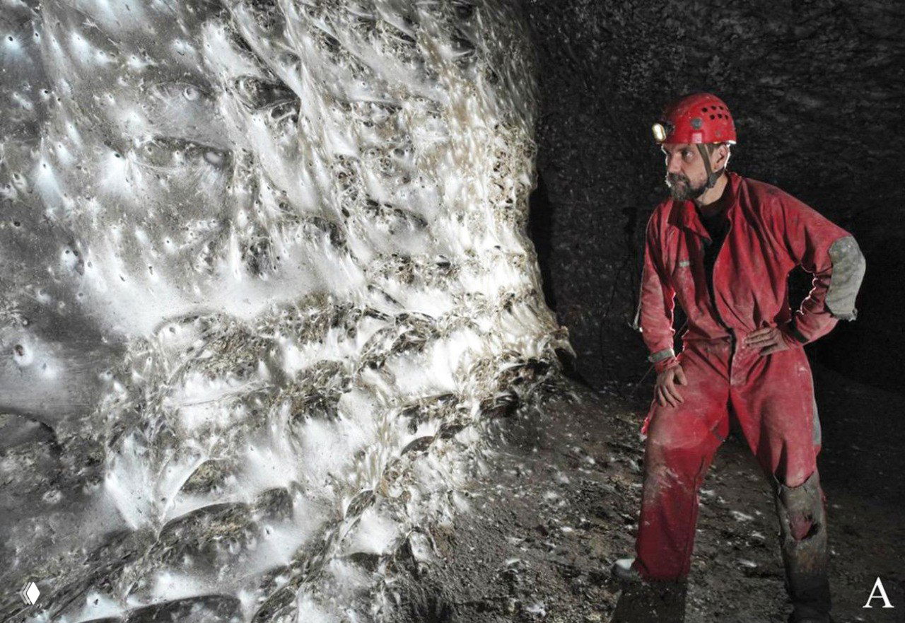 Спелеолог в красном комбинезоне освещает плотный слой паутины на стене Серной пещеры; видна текстура паутины и масштаб по отношению к человеку, около 110 символов