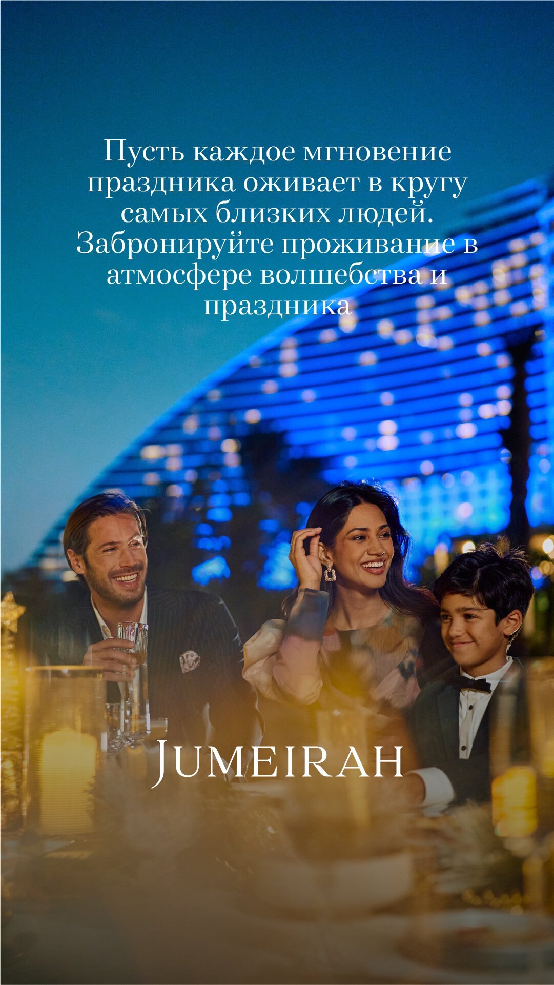 Семья и гости за праздничным столом вечером на фоне подсвеченного здания отеля и праздничной иллюминации — новогодняя атмосфера торжества.