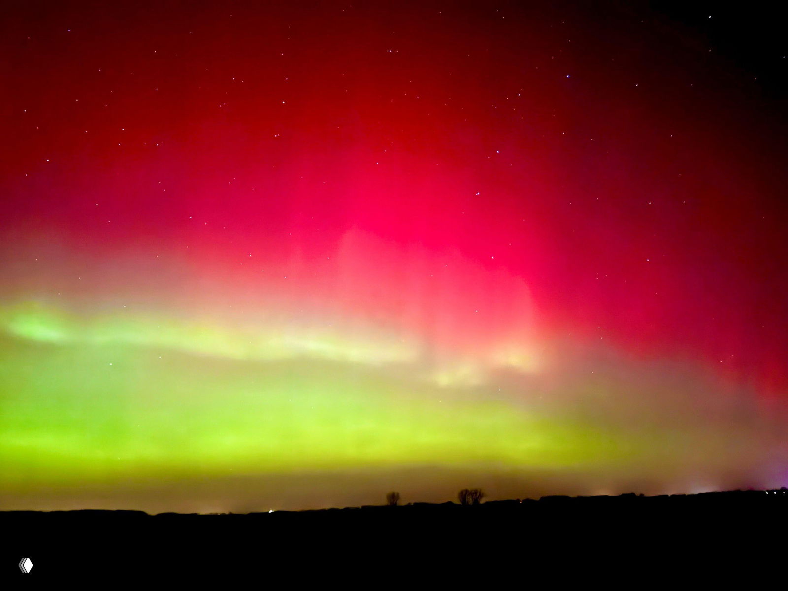 Широкая панорама ало‑зелёного северного сияния, занимающего почти весь кадр; внизу виден тёмный горизонт и слабые огни далёких поселений под ним.