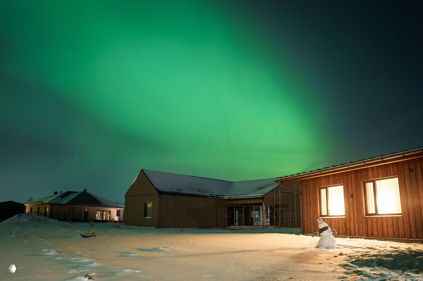 Плотное зелёное полярное сияние, заливающее ночное небо над сельскими постройками; на земле видны следы снега и редкие звёзды над горизонтом.