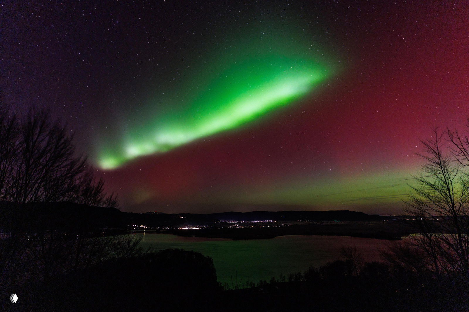 Яркие зелёные и красные полосы сияния над озером и лесом; внизу заметны огни небольшого города и контуры деревьев на берегу ночью.